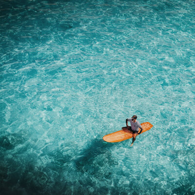 Man sitting on an orange surfboard in turquoise water, symbolising Financial Freedom.