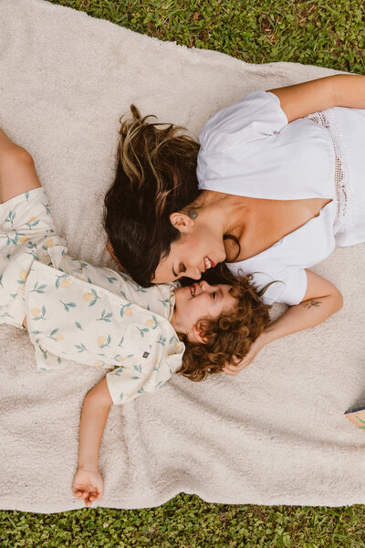 Mother and young son lying on a white blanket in the grass, sharing a playful and joyful moment during their family session at Walter Hall Park in League City, Texas.