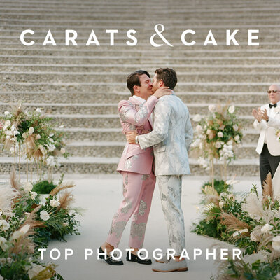 Couple in floral suits kiss on a stone staircase, surrounded by floral arrangements, with a smiling man in sunglasses clapping in the background.