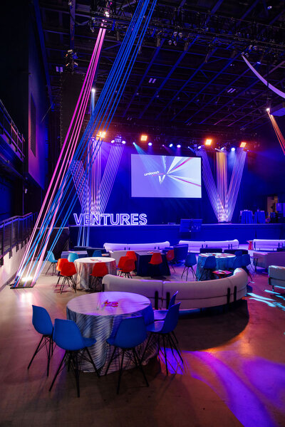 Modern event space with colorful lighting, round tables, and a large screen.