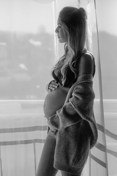 Eine schwangere Frau steht an einem Fenster und schaut raus, während sie ihren Bauch hält.