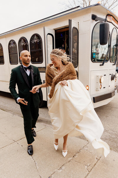 Bride in elegant gown and fur shall holds hands with husband in tux as they step off trolley on wedding day.