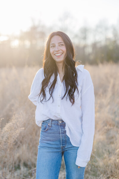 A woman in a white shirt and jeans smiles while standing in a field at sunset