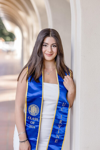 A young, brunette woman leaning against a white wall in an outdoor corridor, wearing a white dress with a dark blue graduation sash that says "UC Irvine," smiling at the camera.