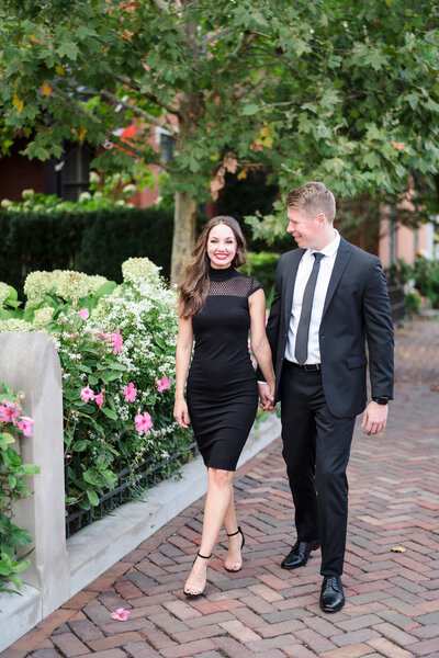 An engaged couple hold hands and walk along the cobblestone sidewalks of German Village with flowers growing in the background