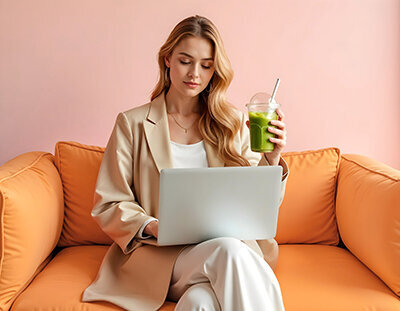 Femme aux longs cheveux blonds, vêtue d'un tailleur beige, assise sur un canapé orange, travaillant sur son ordinateur, matcha glacé à la main.