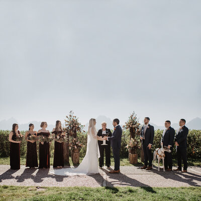jackson hole photographers  captures grand teton wedding  ceremony with bride and groom holding hands and bowing their heads as their officiant says a prayer and their bridal party surrounds them  at jackson hole wedding venues