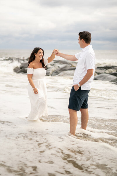 A couple is enjoying a day at the beach. The woman in a white dress holds the hand of the man in a white shirt and dark shorts as they stand in shallow water, against a backdrop of rocks and waves under a cloudy sky.