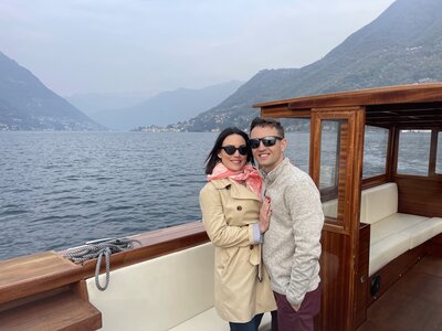 A couple wearing sunglasses stands on a boat. The woman wears a tan trench coat and pink scarf, while the man is in a light sweater. They're embracing, smiling with mountains and a lake in the background under a cloudy sky.