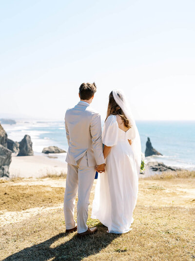 A bride and groom hold hands facing away from the camera with the ocean in the background.