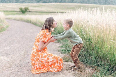 Woman in a bright orange dress bending down to kiss her young son who is giggling with her in a field- photo by Maegan R Photography
