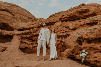 A woman wearing a floor-length white dress holding hands with her fiance who is wearing a tan suit. They are looking in opposite directions to the left and to the right and are standing with beautiful red rocks behind them.