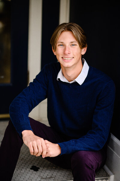 Senior guy outfit ideas with a young man in a dark blue sweater and white collared shirt under and purple pants sitting in a doorway with a smiling face