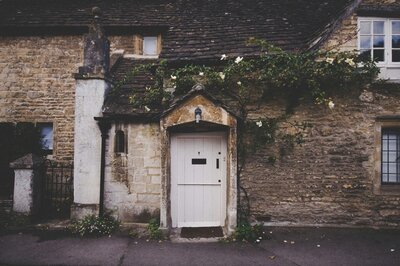 A historic English home crafted from brick and stone, with a front door that opens directly onto the footpath. A climbing vine adorned with blooming roses weaves up the facade, gracefully framing part of the entrance.