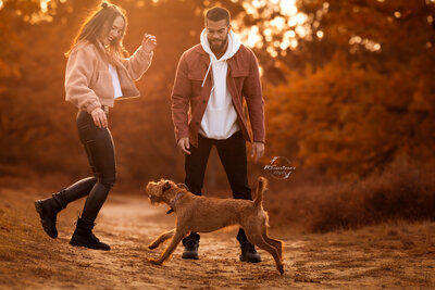 Koppel al spelend met hun hond op een zandpadje tussen de heide tijdens zonsondergang.