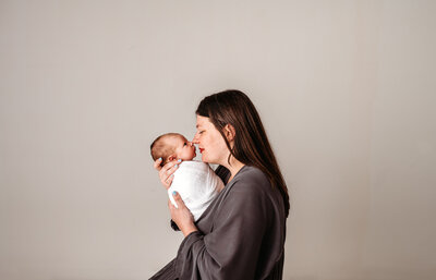 Family photographers Maryland capture newborn pictures with mother in a grey robe holding her newborn baby who is wearing a white swaddle for an in studio session