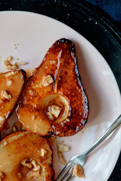 A close-up shot of caramelized pear halves topped with chopped nuts, served on a white plate. Photographed by Mica McCook, this image captures the rich, golden caramelization and the natural sweetness of the pears