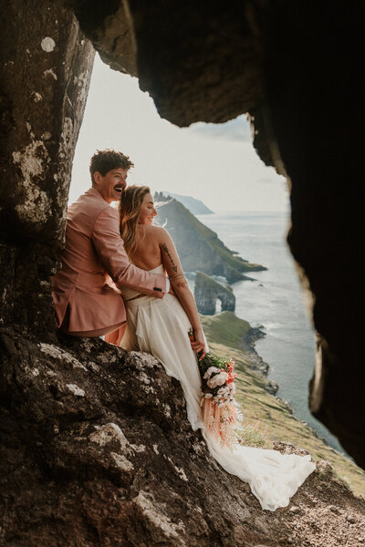 A couple eloping in the Faroe Islands stands at a dramatic viewpoint laughing together