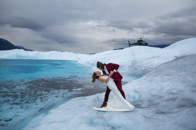 Bride and groom share a dramatic dip kiss on a glacier, with a helicopter in the background under stormy skies.