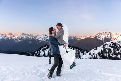 bride is picked up by groom on a snowy ground with skis in the background and snow on ground and pastel skies