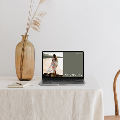 Laptop mockup displaying the Art of Marina website with a flowing floral kimono on a beach, styled in a minimalist interior with neutral decor