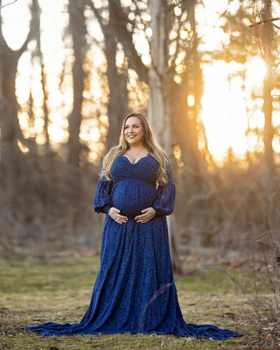A blond woman stands in the forest at sunset, wearing a royal blue lace maternity gown, smiling and holding her belly with both hands.
