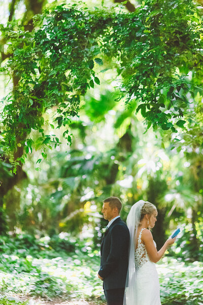 bride and groom stand in the forest reading personal vows to each other standing back to back