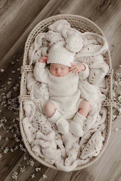 Lehigh Valley Newborn Photographer LG Photography Lori Generose newborn baby in cream outfit and knit booties on a cream blanket in a moses basket with some white flowers around it on a wood floo