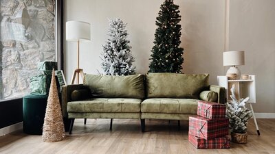 A gray couch with two Christmas trees set up behind it and presents stacked up next to it.