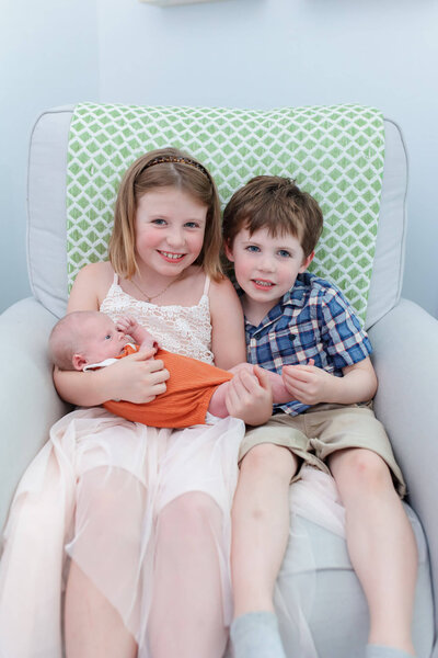A brother and sister holding their newborn brother in a chair in their NOVA home.