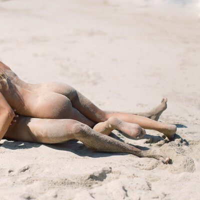 Adventurous couple's boudoir session showing sandy legs and butts intertwined on the beach in Mexico.