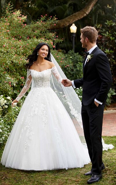 LACE AND TULLE BALLGOWN WITH OFF-SHOULDER SLEEVES Regal, modern, glamorous. Style D3183 from Essense of Australia was made for your grand entrance into wedded bliss! An unexpected neckline style, this subtle, straight-across neckline extends into an off-the-shoulder design with organic lace edging. The long sheer sleeves are wrapped with organic floral laces for an unexpected design detail, which perfectly complements the semi-sheer bodice. The exposed boning and organza layer create that subtle corsetry-inspired look and feel, while the lace motifs extend onto the full ballgown skirt and increase in size toward the hem. Subtle beading is incorporated into the floral appliqués for a rich effect, enhancing the multiple layers of tulle to make this gown the crowning moment of any dream look.