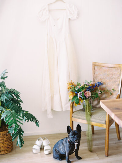 A dog sits for a formal portrait in front of the wedding dress, wedding bride's shoes, and the bouquet, designed by Moonlight Floral Co. for the colorful and festive wedding at Dos Pueblos Orchid Farm in Santa Barbara.