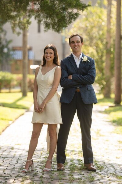 Happy newlywed couple posing for portrait on cobblestone street after St. Augustine courthouse wedding. Bride wearing white sleeveless dress and groom in navy blazer with boutonniere, standing arm-in-arm under dappled sunlight with historic buildings and trees in background.