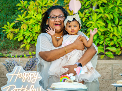 A laughing woman holds a smiling one year old girl in front of her cake on her birthday. Photo by SAVI Photography - San Diego California Photographer