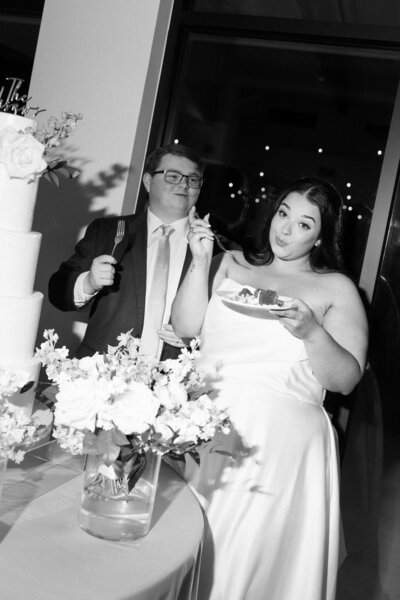 A couple stands by a table with a multi-tiered cake. The woman, wearing a white dress, holds a fork and a plate with cake, smiling. The man, in a suit and glasses, stands beside her holding a fork. A floral arrangement is on the table. Black and white image.
