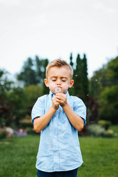 A blonde boy in a blue button-up t-shirt stands in the middle of a garden and blows white dandelion seeds directly at us.