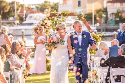 At Palafox Wharf Waterfront Wedding Venue is A joyful wedding scene featuring a bride in a white lace dress holding a bouquet of colorful flowers and a groom in a blue suit, walking hand in hand down an aisle. They are surrounded by bubbles and guests seated on white chairs, with a floral arch and boats visible in the background on Palafox Wharf Waterfront Wedding Venue's downtown waterfront which is a wedding place in Pensacola FL
