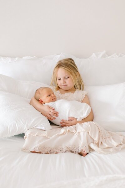 A little girl holds her baby brother while sitting supported on a bed.