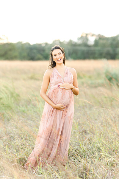 An expecting mother holding her belly and smiling at the camera outside in a field at sunset by northern virginia baby photographer