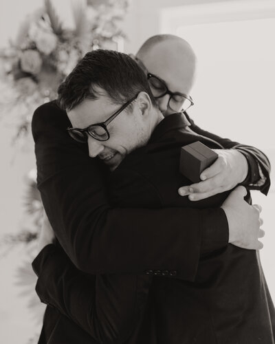 two men embracing in front of floral backdrop while one holds a ring box