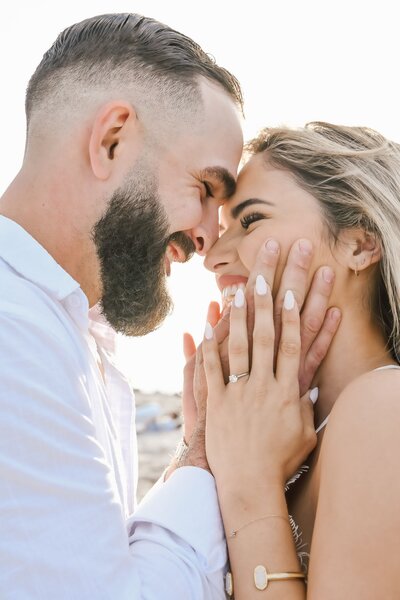 Happy engaged couple sharing a joyful moment on the beach, with the bride-to-be showcasing her soft glam makeup, styled hair, and elegant nails by Blushed Miami.