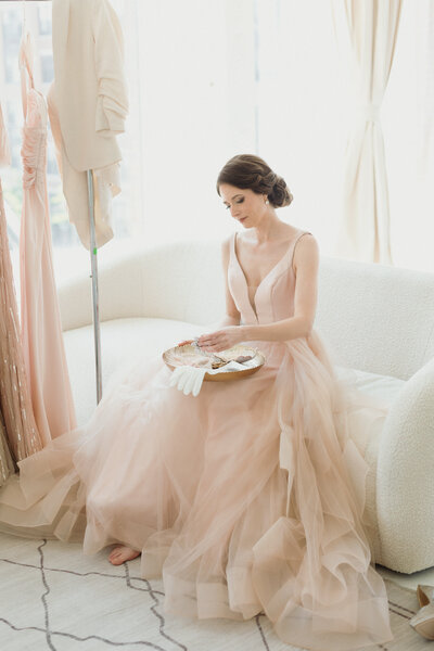 Light skinned woman with dark hair in pink ballgown sitting on couch and looking at her jewelry on a gold tray.