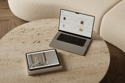 A laptop and iPad rest on a travertine table, displaying a virtual interior styling concept and curated art selections. A boucle sofa and herringbone timber floors add warmth in the background.