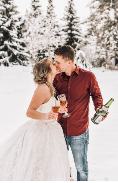 woman in white wedding dress with man in jeans and button down shirt holding champagne glasses outside in the snow