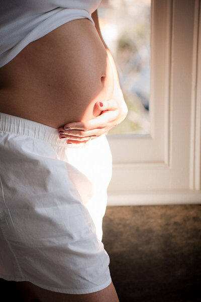 Side view of pregnant belly in white clothing with hand gently resting on abdomen near window in soft natural light.