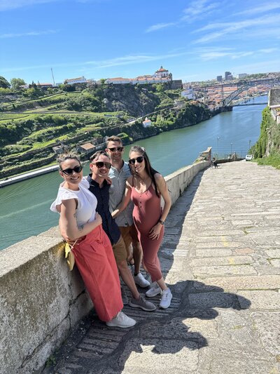 Four people are standing on a stone walkway with a scenic view of a river and lush hills in the background. They are smiling, dressed casually, and posing for the photo in sunny weather. A bridge can be seen in the distance.