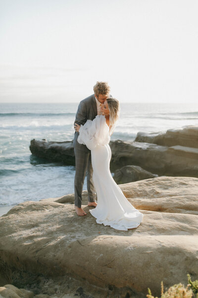 husband kissing his wife on top of rocks by the beach in la jolla