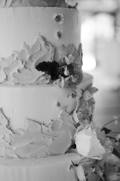 A close up black and white shot of a tiered wedding cake. There's smooth frosting with a layer of a darker textured frosting in waves around the bottom half of each tier. Accented with clusters of fresh flowers.