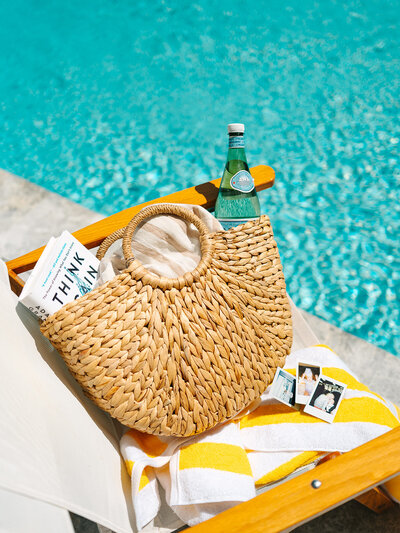 Beach bag filled with items sitting on a yellow striped towel on a chair next to the pool
